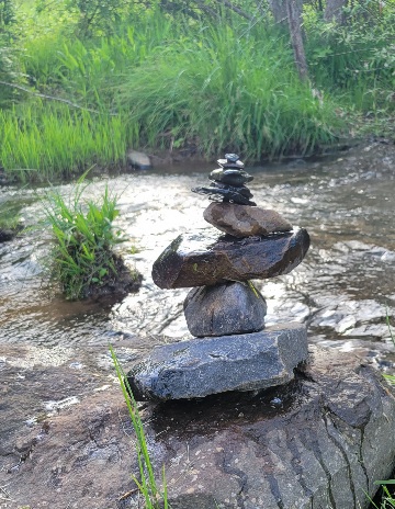 A stack of rocks placed in meditation fashion beside a rippling creek with bright green wild grass in the background.